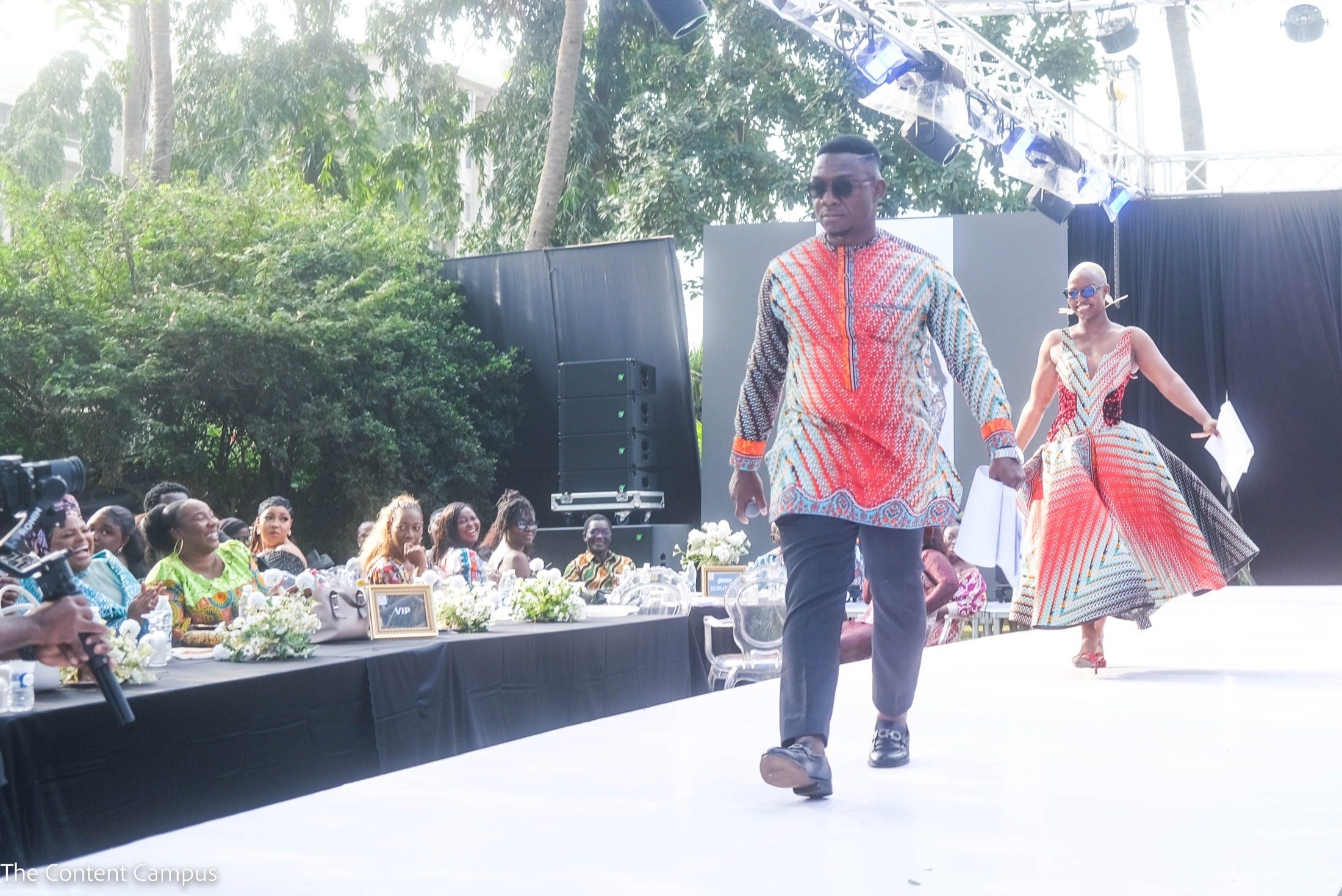 A man in sunglasses and a bright patterned tunic walks along a white runway holding papers, while a smiling woman in a full patterned gown follows behind.