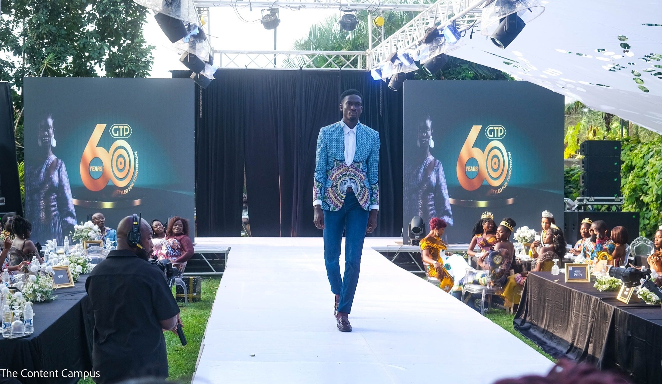 A man in a blue patterned blazer, white shirt and jeans walks down a white outdoor runway as seated guests and a headset-wearing camera operator look on.