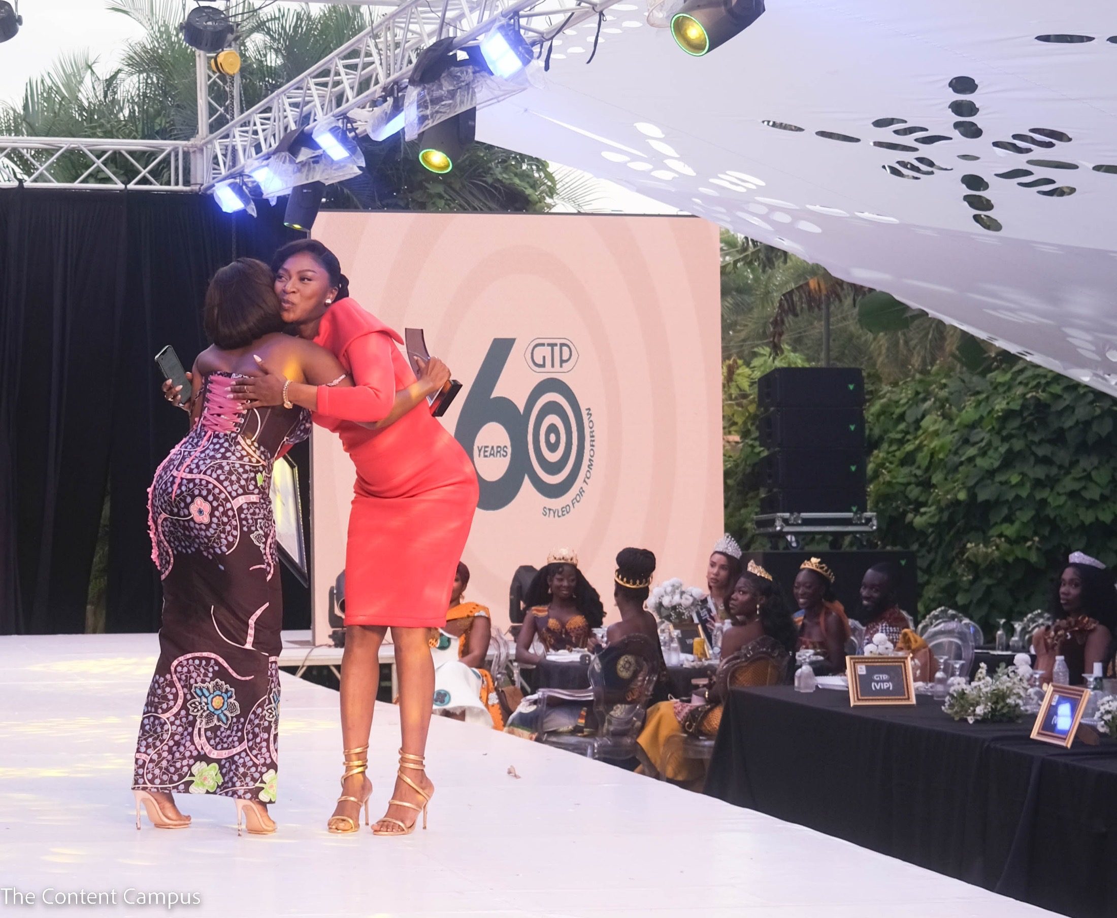 Two women in fitted dresses, one coral and one in a brown print, embrace on a white runway while crowned guests sit at tables behind them.