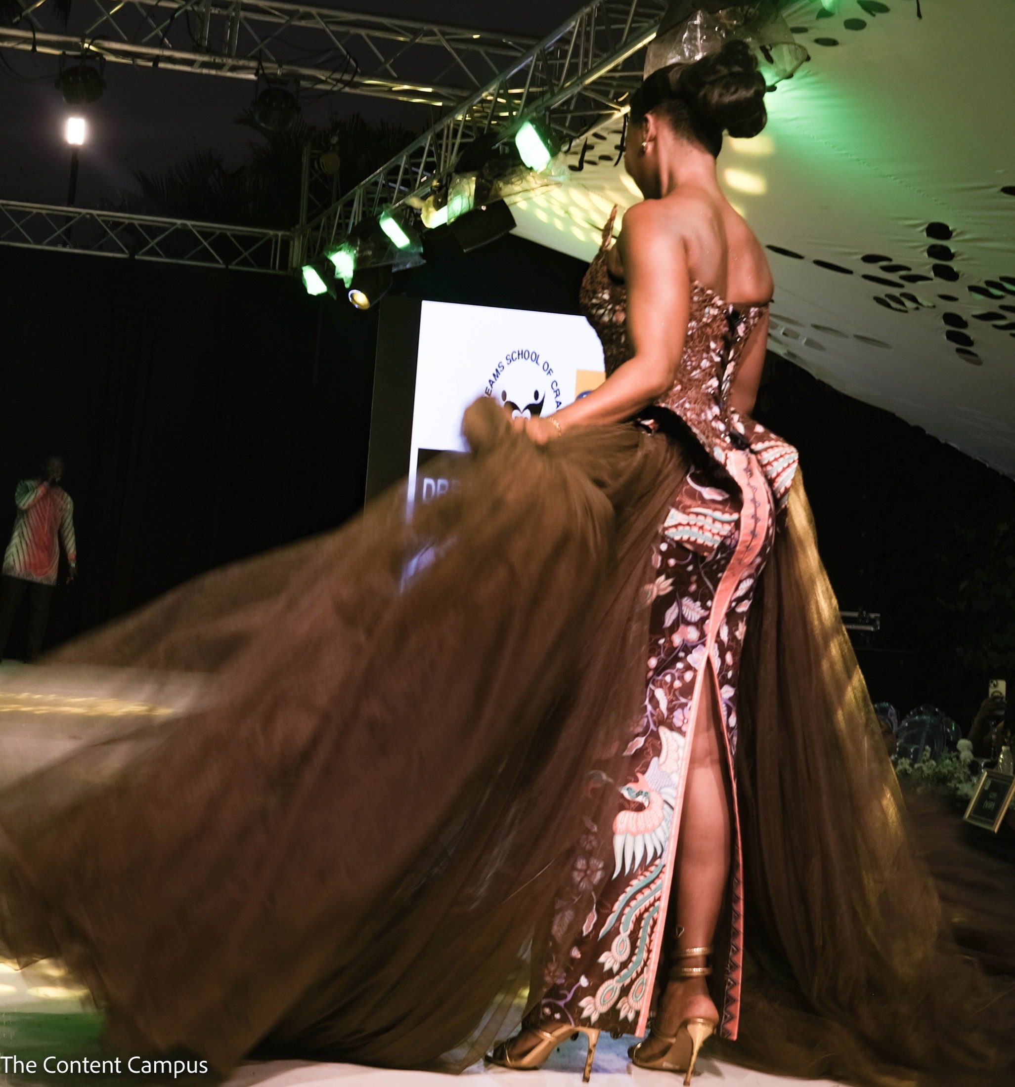 Seen from behind, a woman in a corseted printed gown with a long brown tulle train stands on a runway under green stage lights at night.
