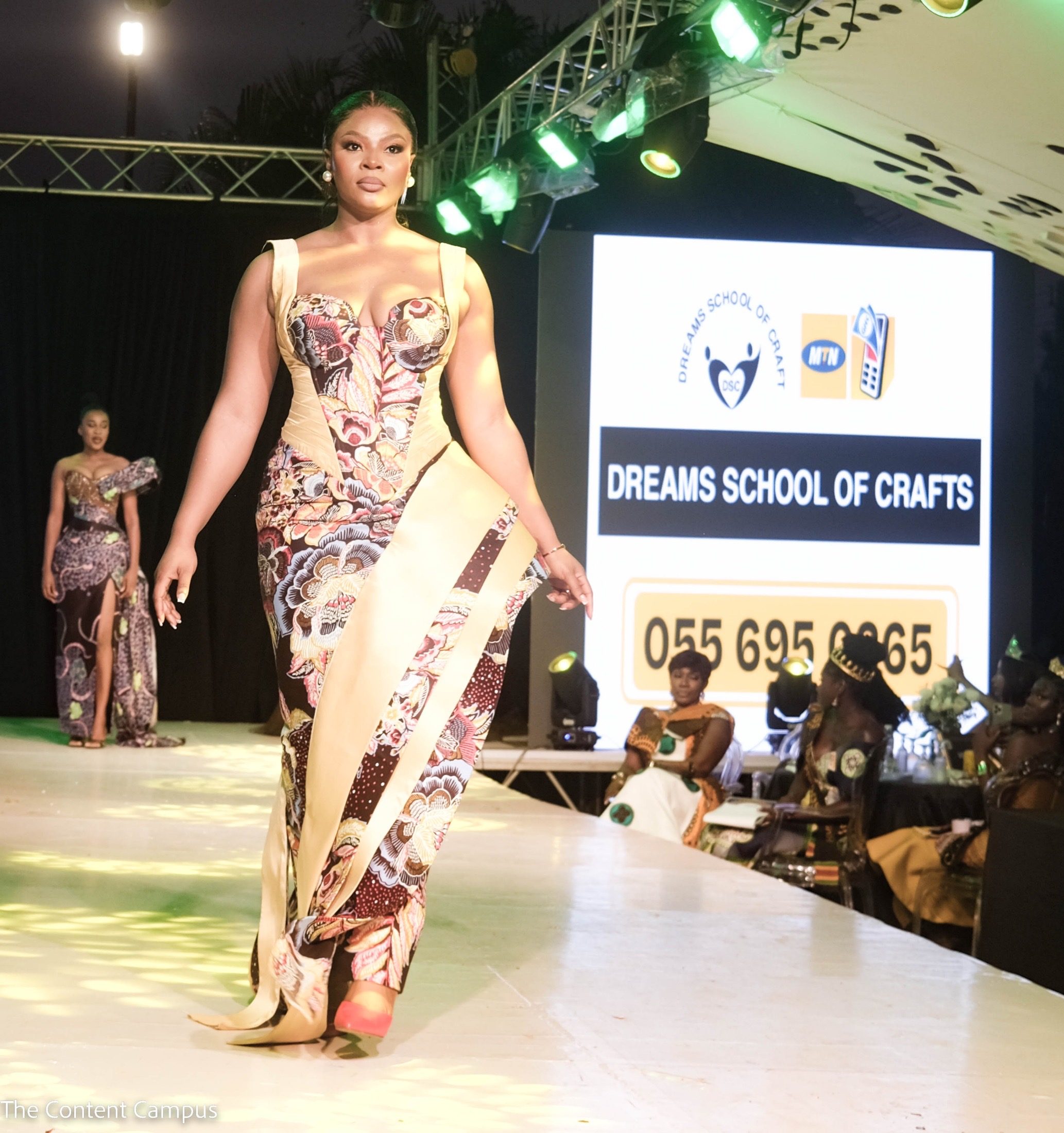 A woman in a floral-print corseted gown with a cream sash walks the lit runway, with another model behind her and a sponsor screen at right.
