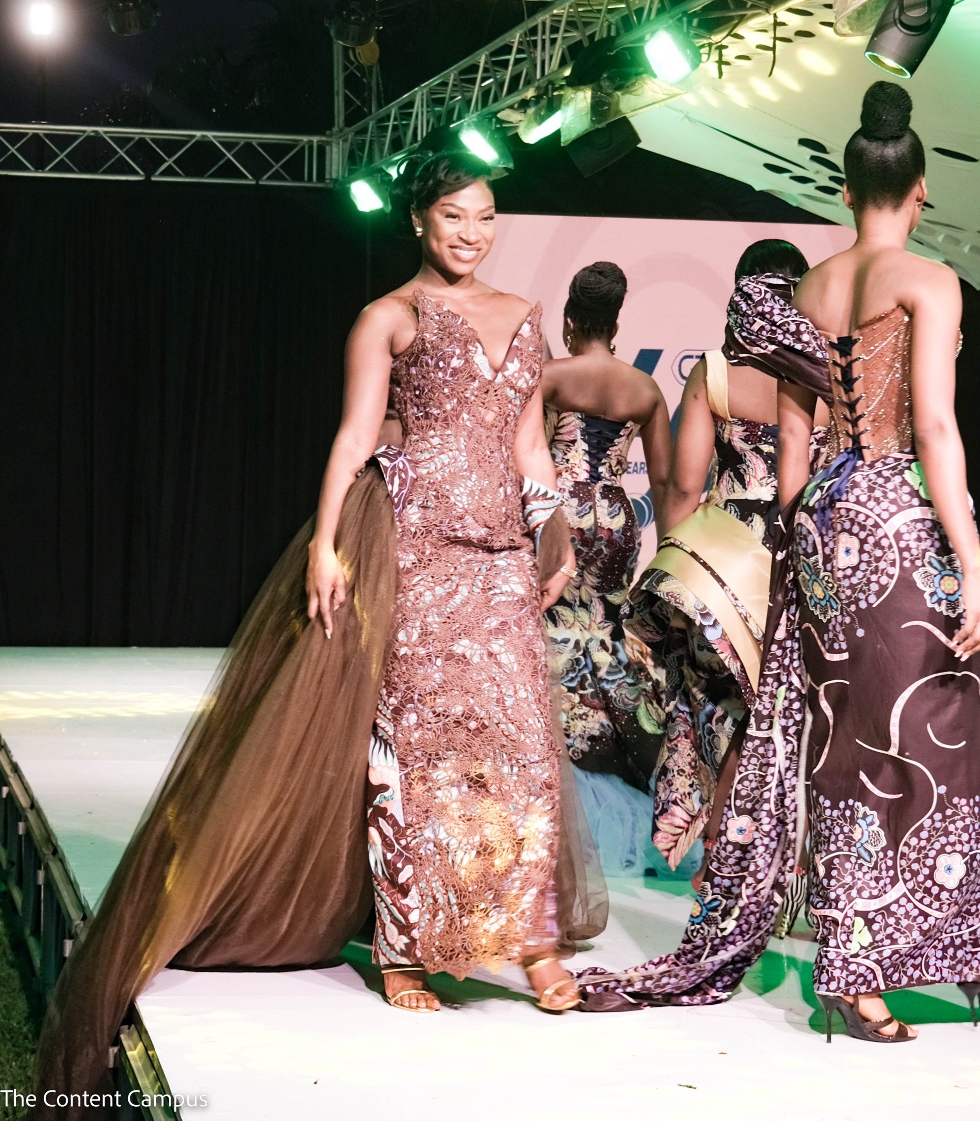 A smiling woman in a brown lace gown with a long tulle train walks forward on a runway while other models in printed gowns walk away.
