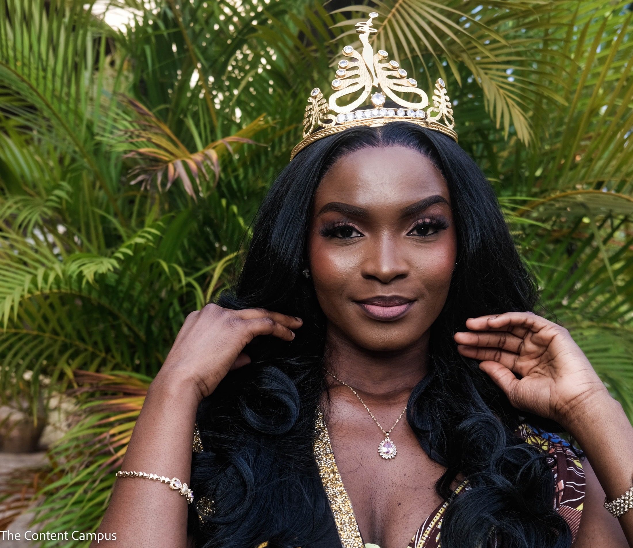 Close-up portrait of a woman with long black wavy hair wearing a gold jeweled crown, pendant necklace and bracelets, with both hands raised before green palm fronds.