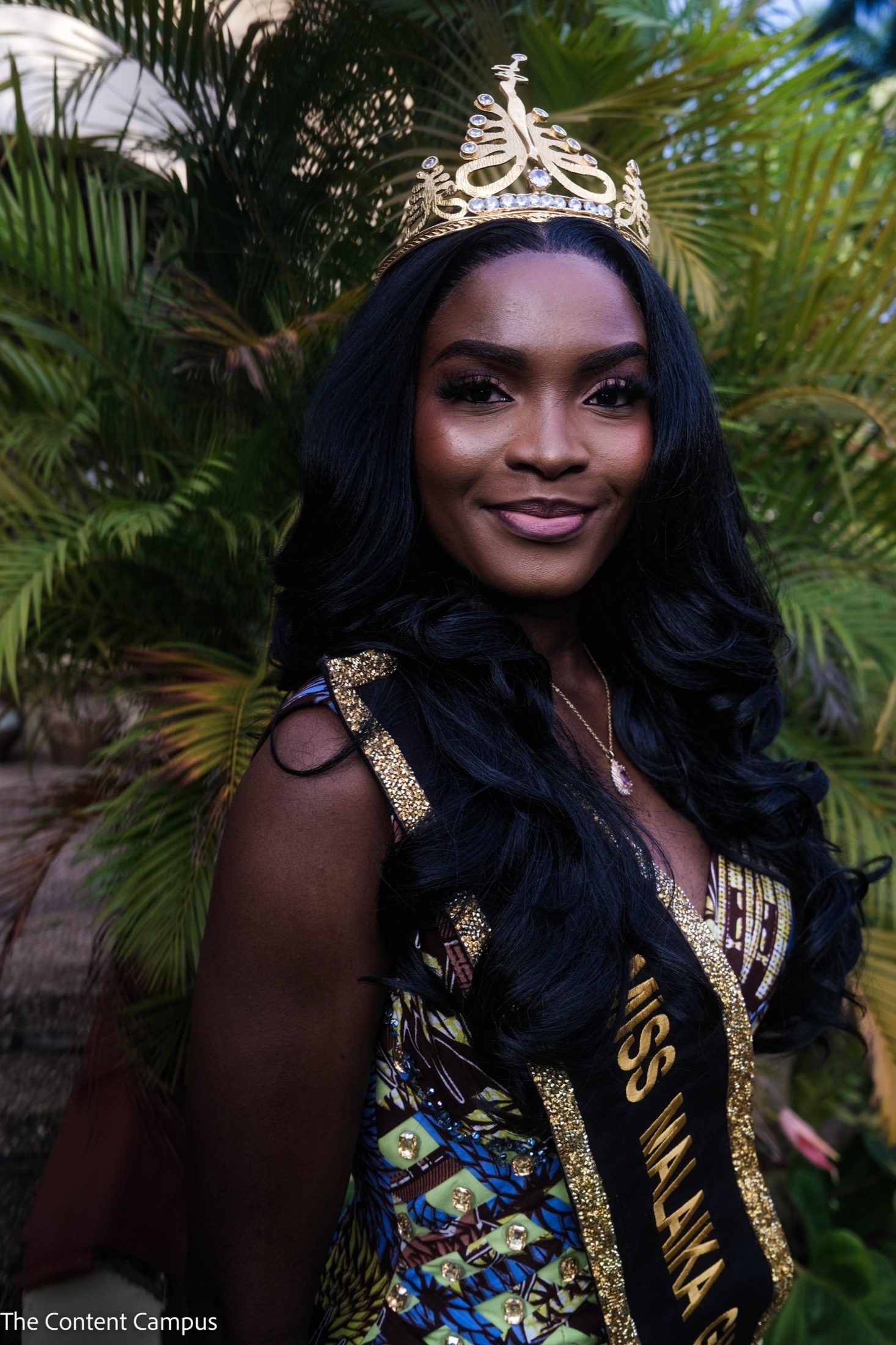 A woman with long, wavy black hair wears a gold jeweled crown, a patterned dress and a black-and-gold sash, standing before green palm fronds in soft light.